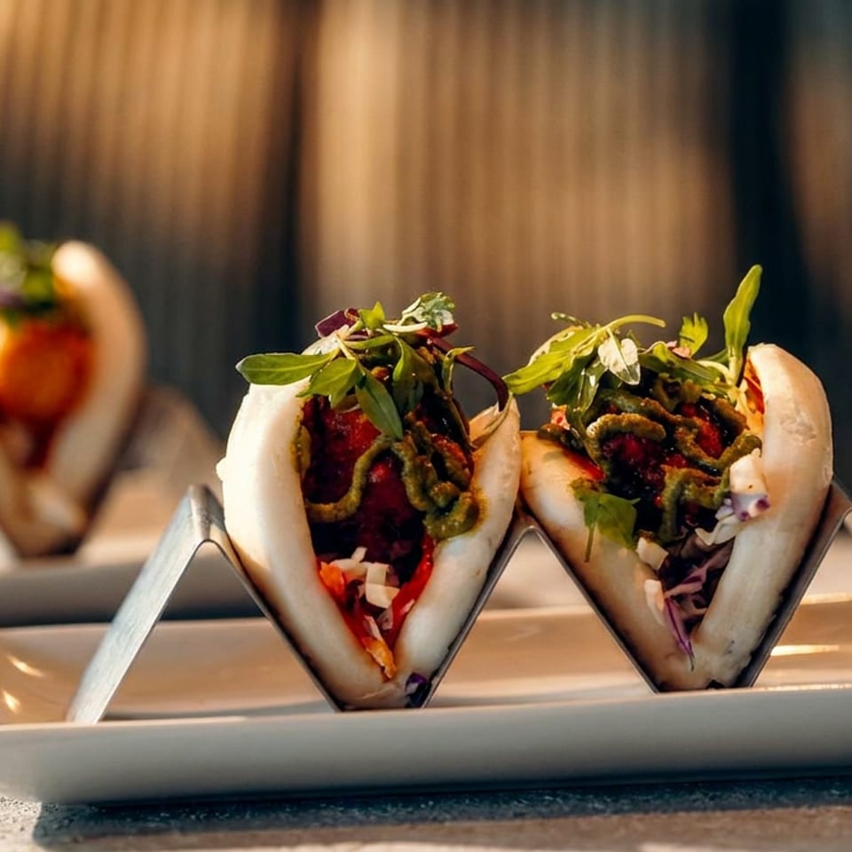 Two steamed bao buns filled with vegetables and sauced meat sit on a white rectangular plate in warm restaurant lighting.