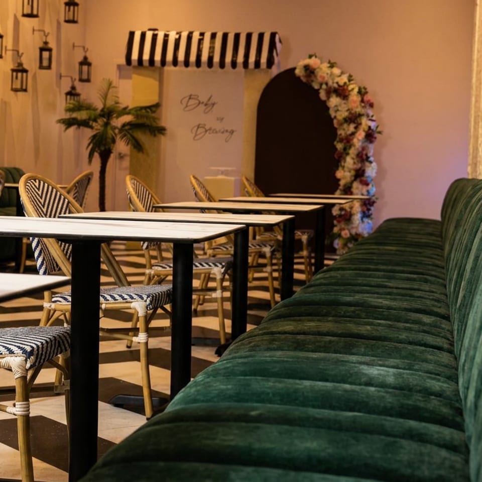 Warmly lit restaurant dining area with green velvet banquette seating, patterned woven chairs, marble tables, and floral archway.